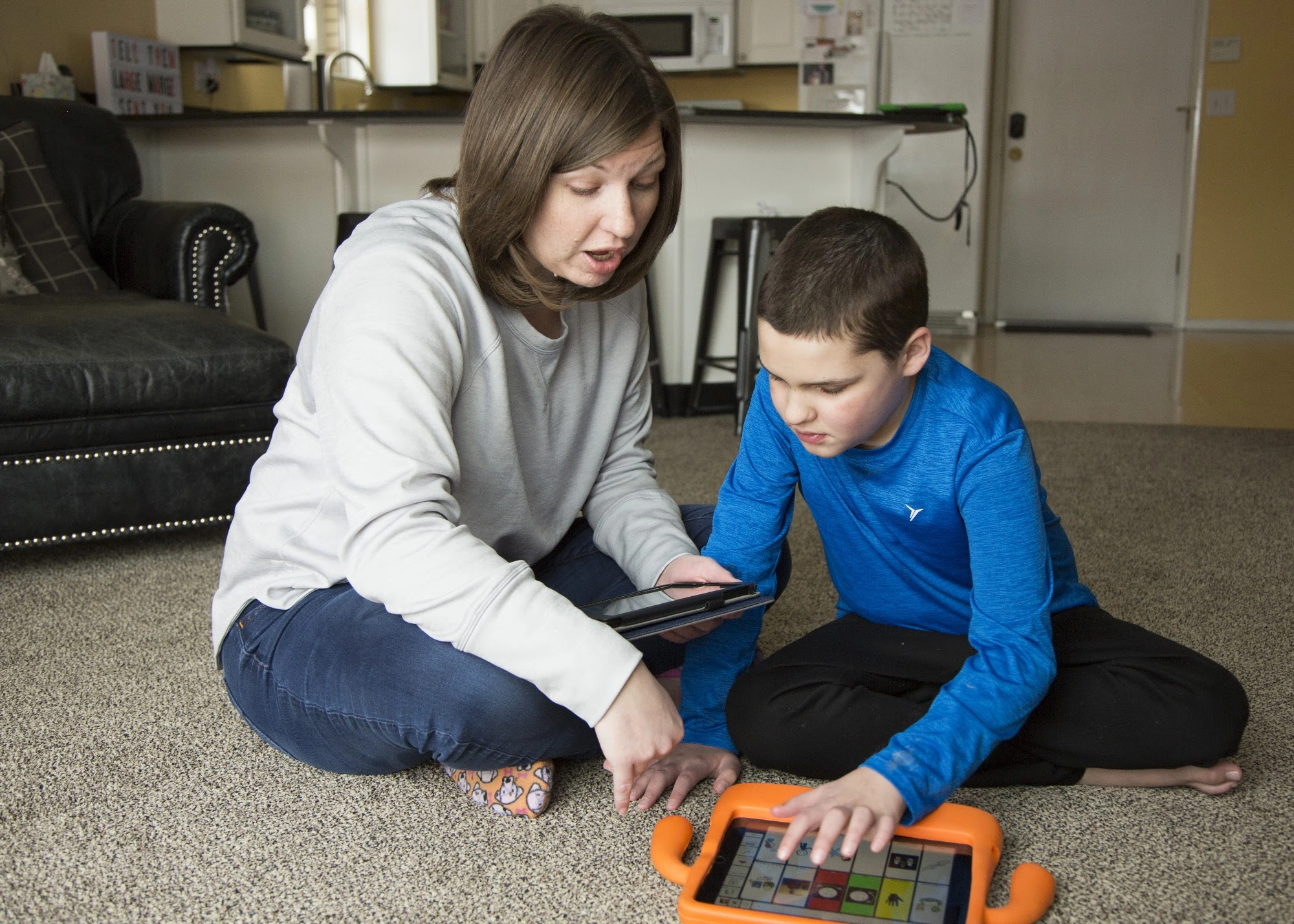 A young AAC user and an adult sit on a rug with a tablet in an orange case.