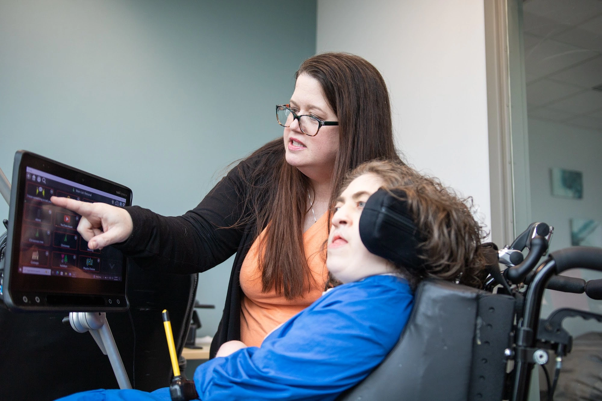 An adult and a wheelchair user communicate using a mounted AAC device in an office.