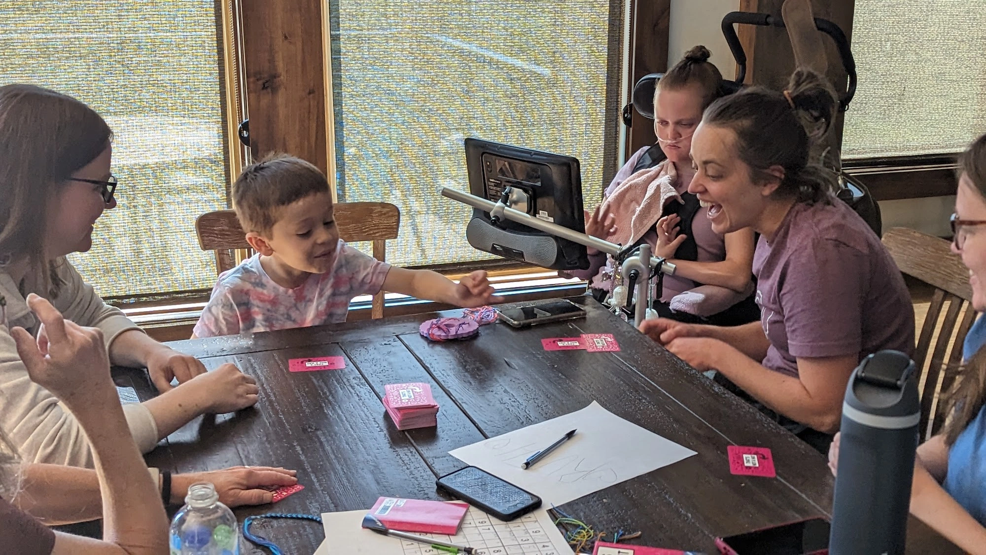 People gathered around a table playing cards, with an AAC device beside a wheelchair user.